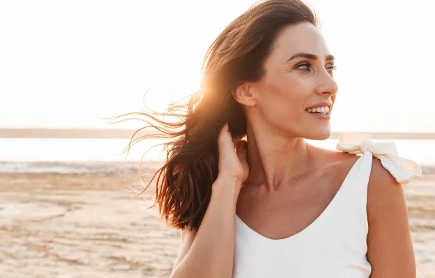 Femme souriante avec des cheveux au vent, soleil en arrière-plan, plage.
